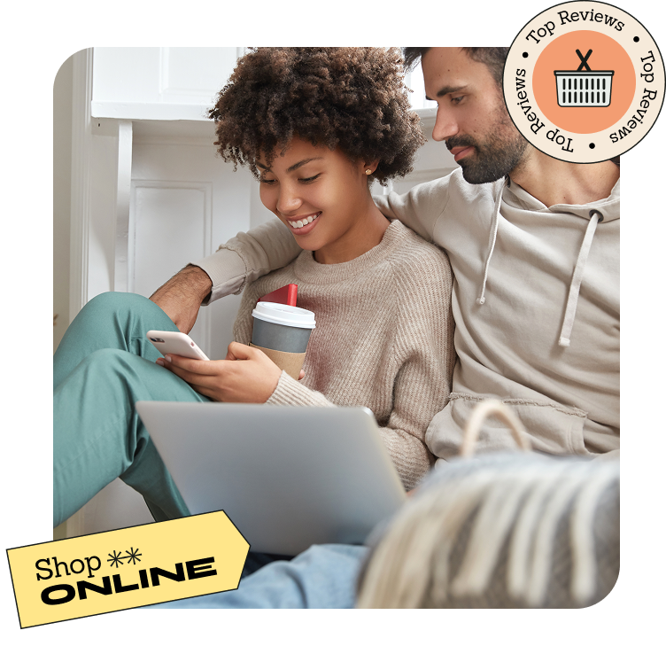 Pareja joven sentada en casa comprando online con móvil y portátil, sonrientes y relajados.
