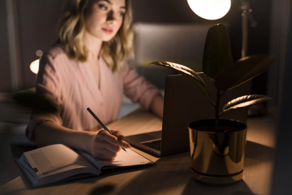 Mujer escribiendo en una libreta junto a su laptop, con la mejor lámpara de lectura led para escritorio.
