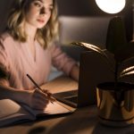 Mujer escribiendo en una libreta junto a su laptop, con la mejor lámpara de lectura led para escritorio.